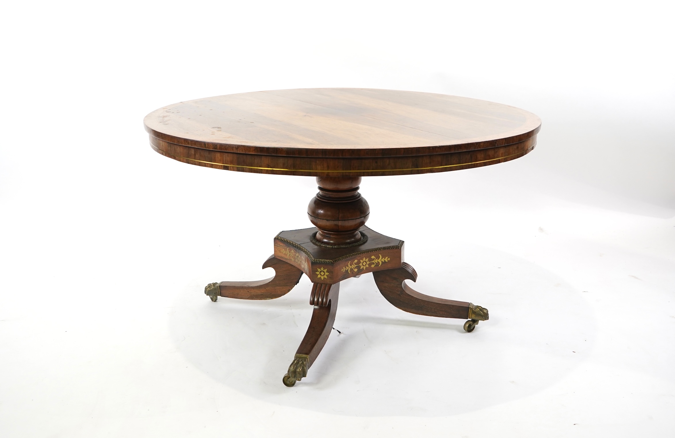 A Regency cut brass inlaid and gilt metal mounted circular rosewood breakfast table, the bird's-eye maple banded tilt top on baluster column, concave platform and quadruple downswept base with cast claw caps and castors,